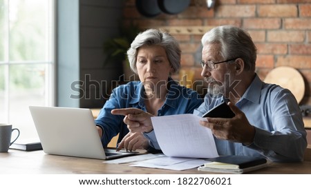 Focused senior husband and wife sit at table at home look at laptop screen pay bills taxes online. Concentrated mature man and woman couple make internet payment on computer, manage finances