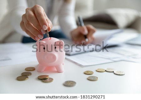 Close up of economical young woman manage household family budget calculate finances at home. Provident female put coin in piggy bank saving invest money for future payment. Investment concept.