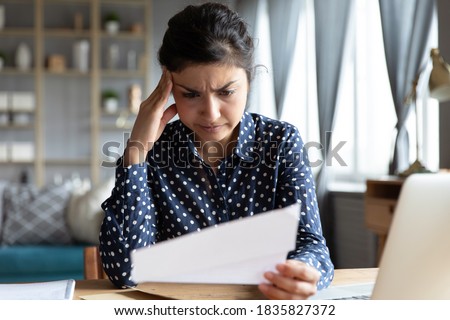 Indian woman sit at desk hold document read paper letter feels disappointed shocked by bad news. Financial trouble bank debt notice, medical test results health problems, huge domestic bills concept