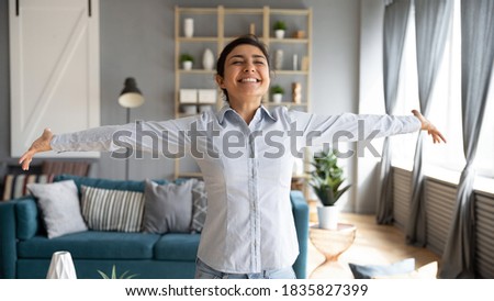 Lively Indian girl having wide smile standing with arms stretched eyes closed enjoy weekend at new house feels free from life troubles, sunlight illuminates cozy room. Welcoming new happy day concept