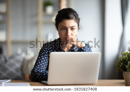 Serious frowning indian ethnicity woman sit at workplace desk looks at laptop screen read e-mail feels concerned. Bored unmotivated tired employee, problems difficulties with app understanding concept 商業照片 © 