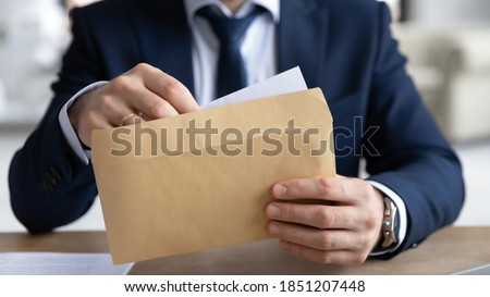Close up young businessman entrepreneur in formal wear opening envelope with paperwork correspondence, male manager employee unpacking banking notification, law order or paper document at workplace.