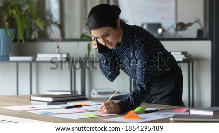 Serious Indian businesswoman talking on cellphone, writing notes on colorful sticky papers in office, employee checking financial documents, working with statistics, consulting client by phone call