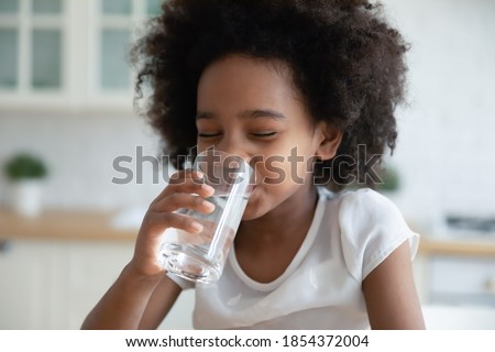 Pretty little African American girl drinking fresh water in kitchen close up, cute preschool child kid holding glass of pure mineral water, enjoying, healthy lifestyle and refreshment concept