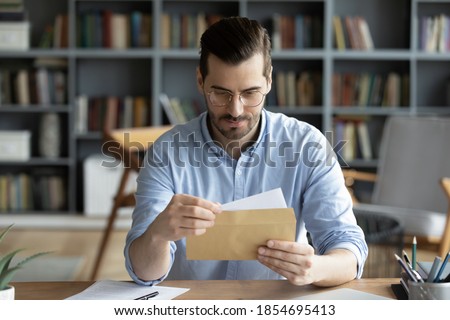 Serious businessman sit at workplace office desk holding envelope take out letter feels interested read business news, got invitation, learns bank statement information. Postal correspondence concept