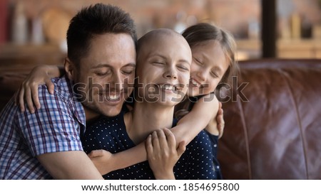 Loving and caring. Affectionate happy young husband dad and school age daughter kid sitting on couch supporting cuddling warm tight beloved smiling millennial wife mom fighting against cancer disease