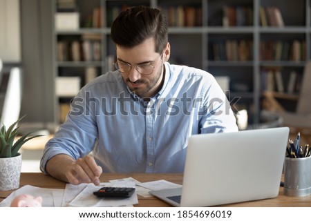 Serious man in glasses sit at workplace desk in office use calculator calculates monthly expenses, taxes, check bank account balance, summarize total sum. Family or personal budget management concept