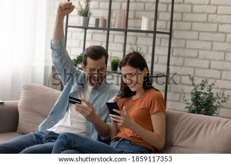 Happy young married family couple involved in online shopping, celebrating getting gift or winning prize in giveaway, paying for goods or services with credit bank card in smartphone application.