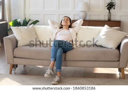 Calm and positive. Happy serene young female relaxing on couch at living room leaning back on pillows. Woman taking break of household chores, breathing fresh air, dreaming about spending weekend
