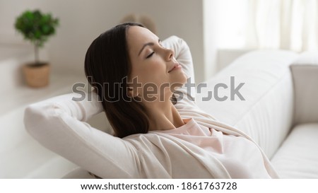 Weekend at least! Tranquil peaceful millennial female enjoying freedom dreaming relaxing on couch with closed eyes, hands behind head. Woman meditating breathing fresh air feeling happy
