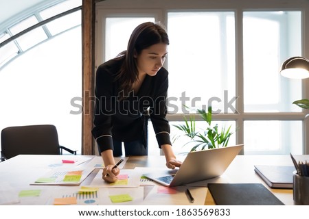Confident millennial indian female business analyst financial advisor preparing statistic report studying documents on work desk, browsing information online using pc, writing out notes on paper sheet Сток-фото © 