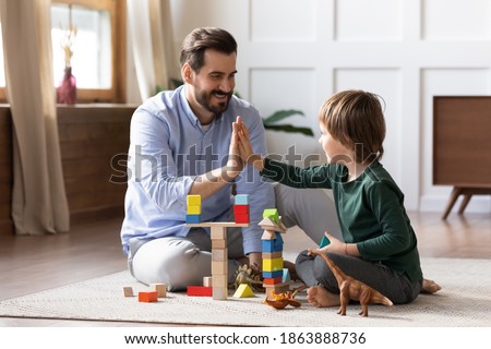Loving young Caucasian dad sit on floor play with small son give high five celebrate success. Happy caring father engaged in activity game build with blocks bricks with little boy child together.