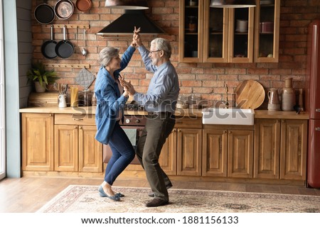 Full length energetic middle aged family couple dancing to disco music in kitchen. Happy old mature man and woman having fun, entertaining together indoors, involved in funny domestic activity.