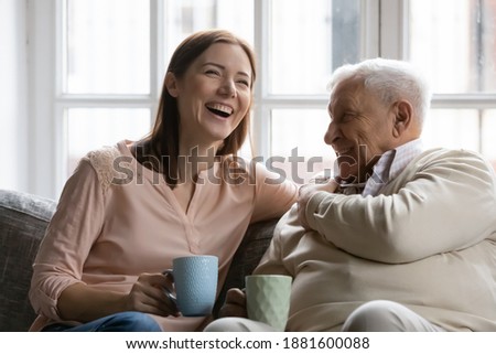 Family members of two generations grown daughter and mature father having fun enjoy tea talk on cozy couch. Attentive young lady caregiver social worker visit support care for glad elderly man patient