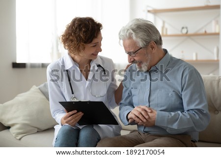 Happy young female doctor nurse laughing, sharing good health test news with happy older senior man in eyeglasses, visiting retired patient at home, medical insurance, domestic geriatric help concept.