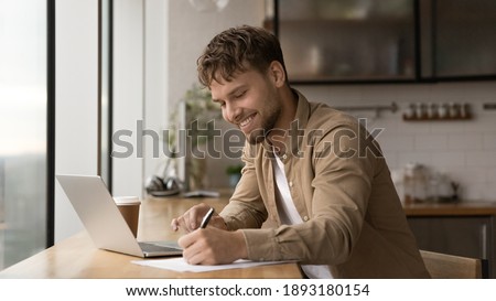 Wide banner panoramic view of smiling young Caucasian man sit at desk make notes working online on computer. Happy millennial male handwrite study distant on laptop in office. Education concept.
