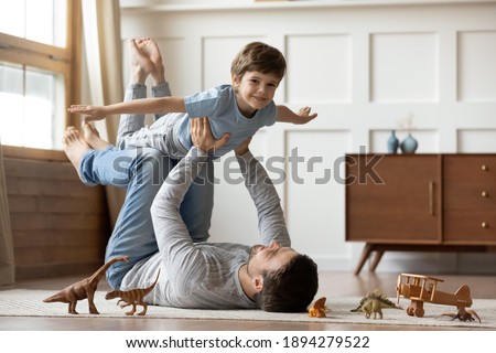 Portrait of happy young Caucasian father lying on floor at home play with cute little 6s son. Loving dad feel playful engaged in funny game activity, hold in hands fly with excited small boy child.