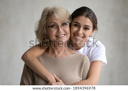 Head shot portrait loving grown up daughter hugging middle aged mother from back, looking at camera, happy mature grandmother and granddaughter posing for family photo on grey background together