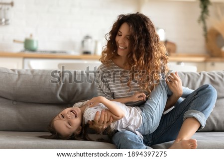 Happy young attractive mother holding on hands cute smiling daughter sitting on couch at home. Close up relaxing parent having fun free time and playing with preschool child.