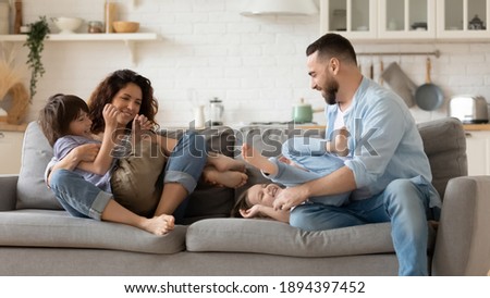 Happy father and mother tickling young daughter and son sitting on couch in living room. Family having fun at home. Parents playing with cute daughter and son