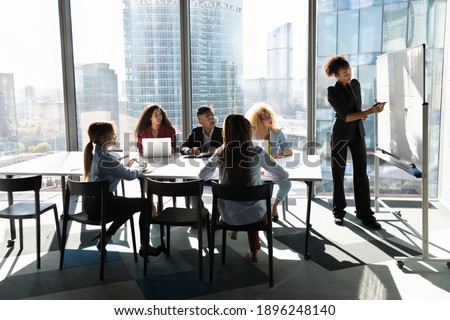 Young African American female coach or speaker make flip chart presentation to diverse businesspeople at meeting in office. Woman tutor or trainer present project on whiteboard to diverse colleagues.