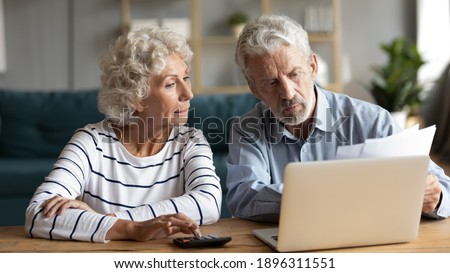 Focused middle aged retired family couple managing monthly budget, involved in financial paperwork, paying taxes online using e-banking computer application or calculating expenses together at home.