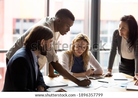 Concentrated young african american employee analyzing marketing research results or sales statistics data at briefing meeting with motivated older korean and young caucasian colleagues in office.