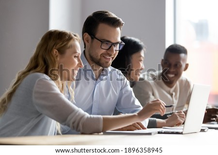 Happy young female employee discussing online project, showing computer presentation to skilled team leader in eyeglasses. Friendly diverse colleagues working in pairs on laptop, using applications.