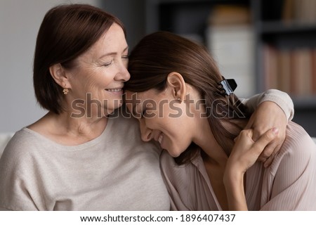 Close up smiling young woman and mature mother hugging, elderly mum and grownup daughter enjoying tender moment, cuddling, holding hands, happy family spending leisure time at home together