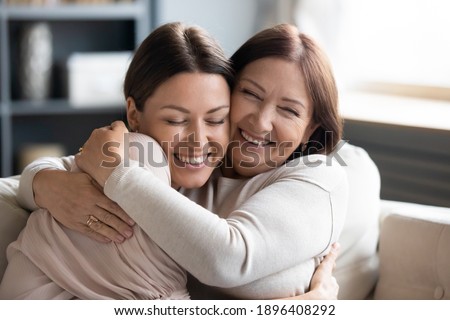 Close up smiling grownup daughter and mature mother cuddling, enjoying tender moment, hugging, expressing love, having fun, sitting on cozy couch at home, family spending leisure time together