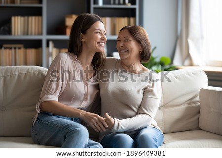 Overjoyed young woman with mature mother chatting, laughing at joke, enjoying pleasant conversation, middle aged mum and grownup daughter hugging, having fun, sitting on cozy couch at home