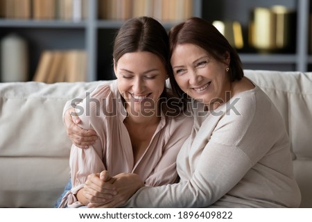 Close up smiling mature mother embracing grownup daughter, having fun, sitting on cozy couch at home, happy young woman with elderly mum enjoying leisure time together, excited by good news