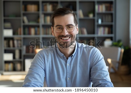 Head shot portrait of happy young handsome confident man in eyeglasses, looking at camera sitting at table at home office. Smiling pleasant businessman holding video call conversation at workplace.