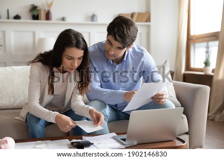 Close up young couple checking mortgage or loan agreement, financial documents together, using laptop and calculator, sitting on couch at home, focused wife and husband calculating domestic bills