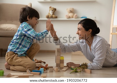 We are great team. Laughing indian mommy give high five to happy kid son celebrate building of high brick tower. Female babysitter construct from brick set with small boy on floor with heating system