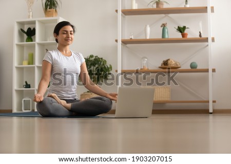 Serene millennial mixed race woman engaged in yoga practice breath deep feel comfort harmony. Tranquil inspired young indian lady sit by laptop on floor at lotus asana meditate to reach peace of mind