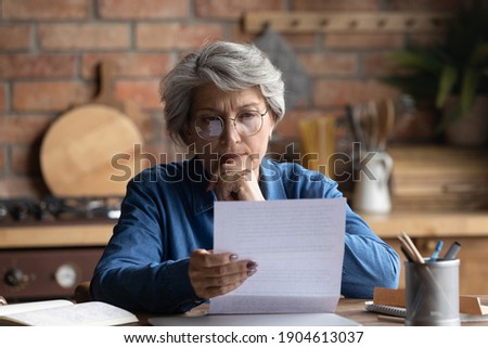 Thoughtful middle aged 60s woman in eyeglasses reading paper letter carefully. Pensive focused older mature retired lady looking at correspondence sheet. considering bank notification at home.