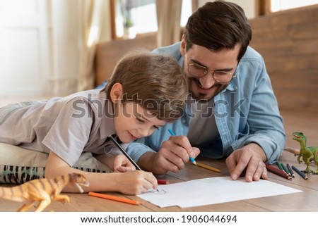 Loving young Caucasian father lying on floor at home have fun painting with little 7s son together. Happy playful dad drawing in album with excited small boy child on weekend at home. Hobby concept.