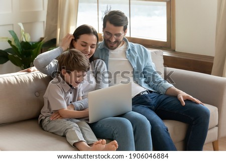 Smiling parents relax on sofa in living room with small 7s son child have fun browsing using modern laptop together. Happy young Caucasian family with little boy kid watch funny video on computer.