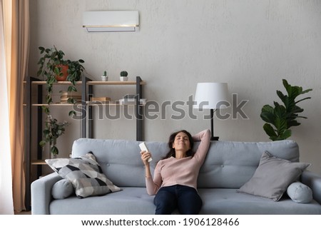Happy young caucasian woman turning on air conditioner with controller, breathing fresh cooled air or enjoying comfortable temperature indoors, relaxing on comfortable sofa in modern living room. Stock photo © 