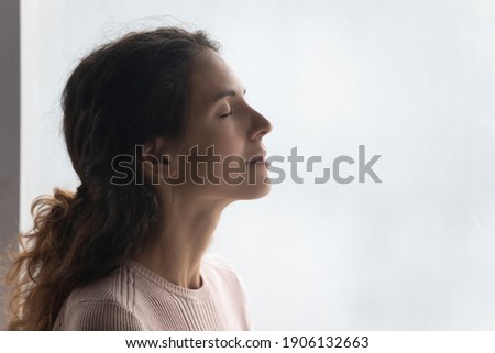 Side head shot view mindful smiling beautiful young woman breathing fresh air, standing near window. Happy millennial caucasian lady enjoying meditation moment, feeling peaceful indoors, copy space.