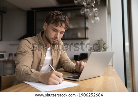 Young guy student prepare research essay at modern kitchen surf internet use laptop write up information to paper report by hand. Focused man freelancer work from home make list of data from pc screen
