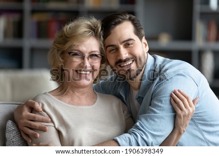 Head shot portrait of smiling young caucasian man embracing affectionate older senior mother in glasses, enjoying sweet tender time indoor, spending weekend leisure together, family relations concept.