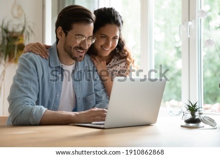 Cheering spouses having warm relations hug by desk at home make shopping purchases online at web store. Friendly young couple in love look at computer screen planning wedding choosing tour on vacation