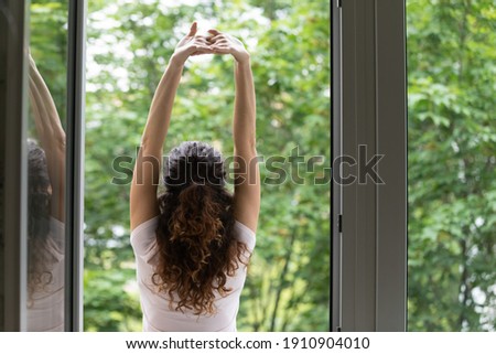 Meeting summer day. Back rear view of happy young woman stand in balcony door stretch hands enjoy weekend have pleasure. Calm beautiful female do morning exercises practice yoga by opened large window