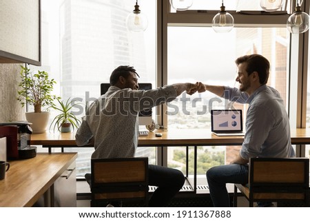 Well done, buddy. Motivated diverse young men coworkers bump fists on workplace feel excited achieve common goal. Two workers international business team members share success glad to help one another