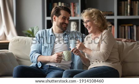 Emotional happy young handsome man telling funny joke or story to laughing middle aged 60s mother, relaxing on comfortable couch with cups of tea, family communication trustful relations concept.