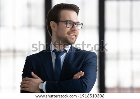 Smiling young businessman in suit and glasses look in window distance thinking or planning career success. Happy Caucasian male director or CEO thinking visualizing in office. Business vision concept.