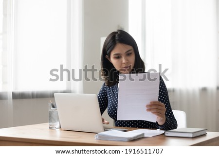 Attentive millennial asian female holding paper letter document reading financial statement. Focused vietnamese business woman worker employee review offer proposal study job contract terms conditions
