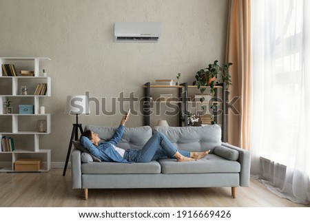 Glad millennial female buyer of air conditioner relax on sofa hold controller breath cool fresh air despite of hot summer day outside. Happy young lady regulate climate at home using modern ac device